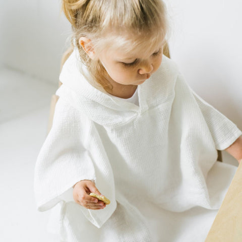2. Child sitting in white linen waffle poncho holding a snack in a bright room