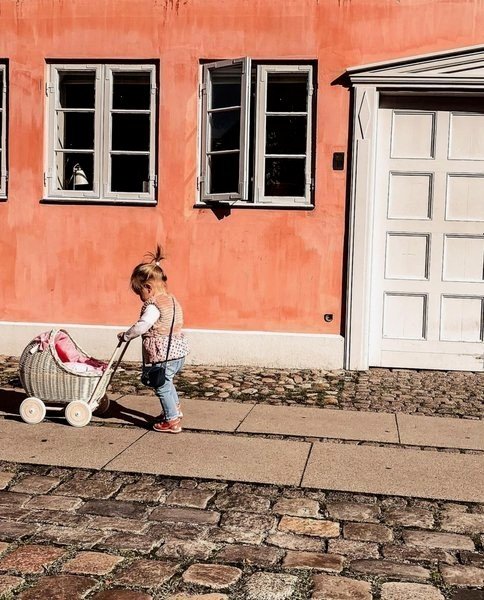 1. Child pushing whitewashed wicker doll pram with beige bedding on cobblestone street