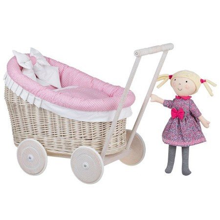 2. Whitewashed wicker doll pram with pink bedding next to a doll in a dress, studio setting