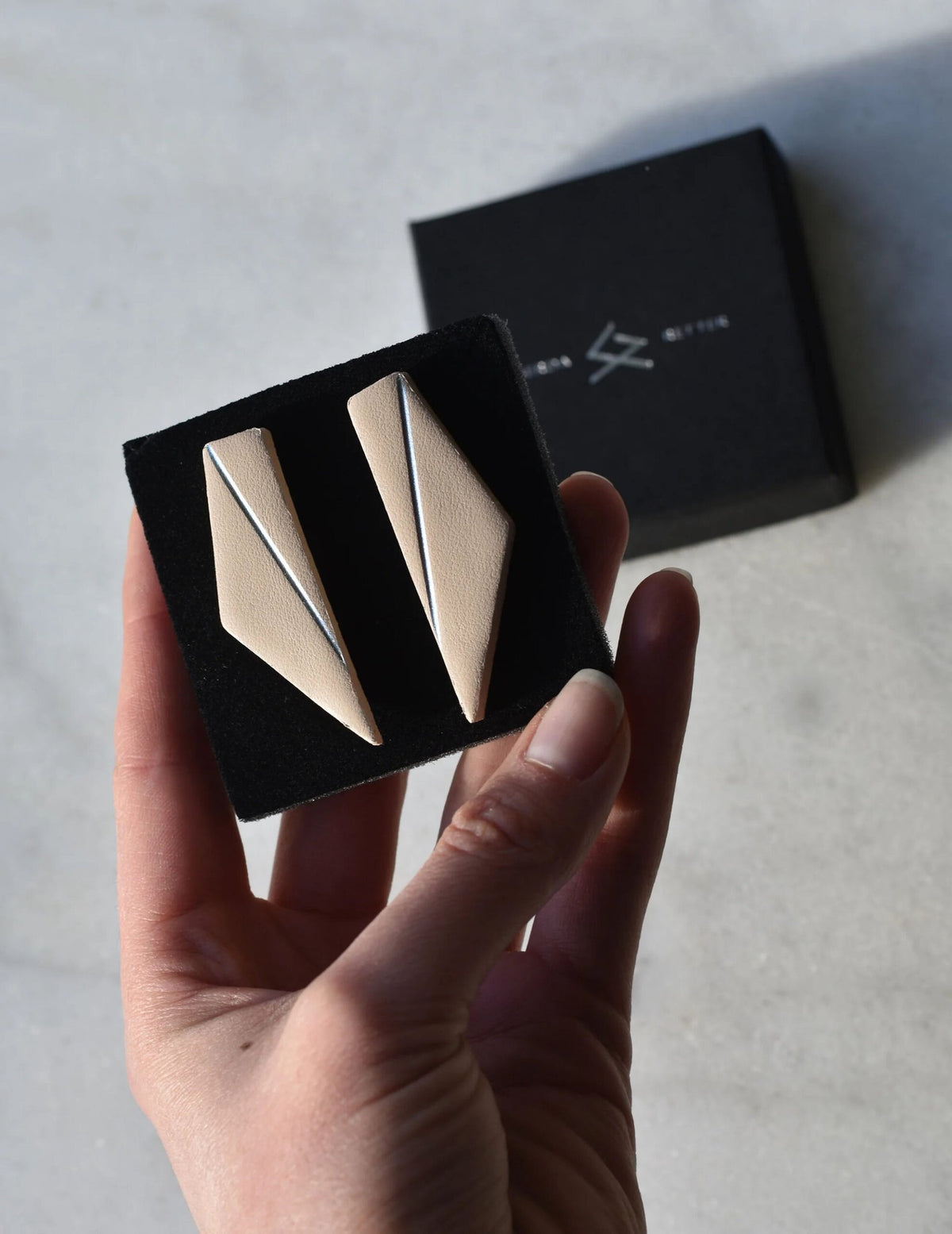 3. Beige Gerda Retter Design Short Wings Earrings in hand, with branded box in background