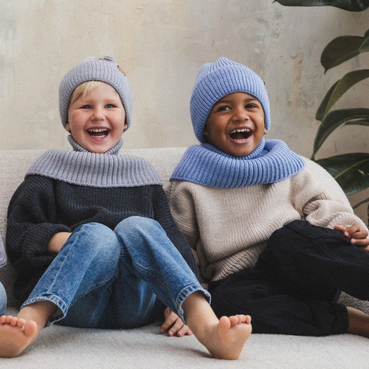 1. Two children laughing, wearing sky blue and grey merino wool hats and neck warmers, sitting on couch indoors