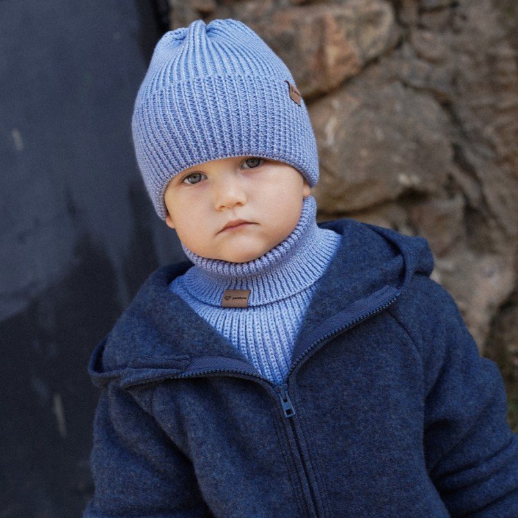 1. Child in sky blue merino wool hat and neck warmer, wearing dark blue jacket, standing outdoors against stone wall
