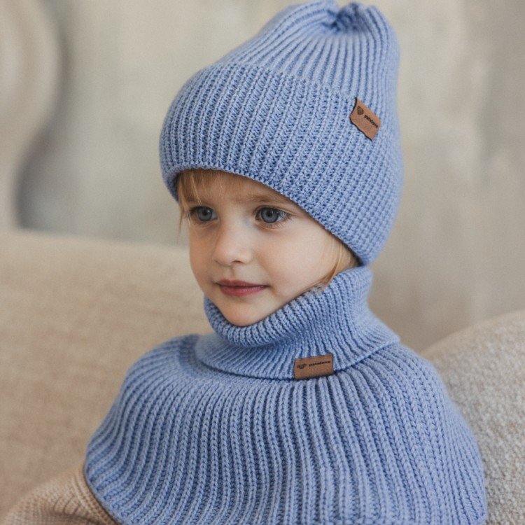 2. Child wearing sky blue merino wool hat and neck warmer, sitting on beige couch indoors