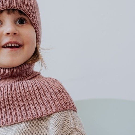 1. Smiling child wearing blush merino wool tube scarf and hat, sitting against a light background