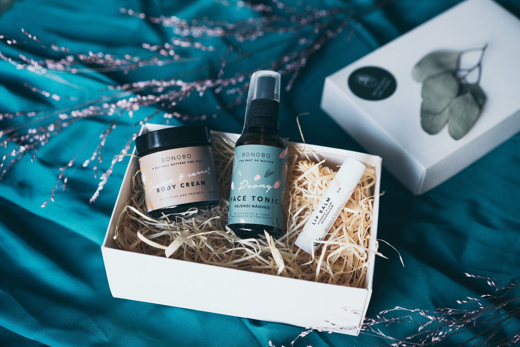 1. Open BONOBO gift box on teal fabric showing Lady’s Mantle Face Tonic, Orange Body Cream, and Avocado Lip Balm nestled in straw