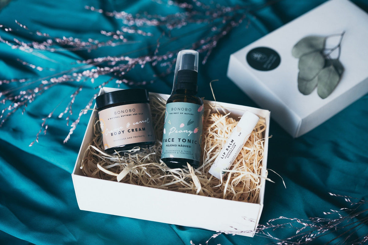 1. Open BONOBO gift box on teal fabric showing Lady’s Mantle Face Tonic, Orange Body Cream, and Avocado Lip Balm nestled in straw