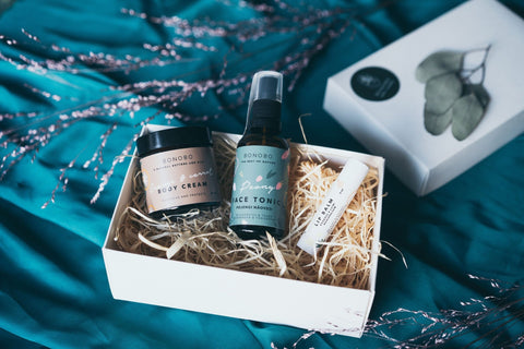 1. Open BONOBO gift box on teal fabric showing Lady’s Mantle Face Tonic, Orange Body Cream, and Avocado Lip Balm nestled in straw
