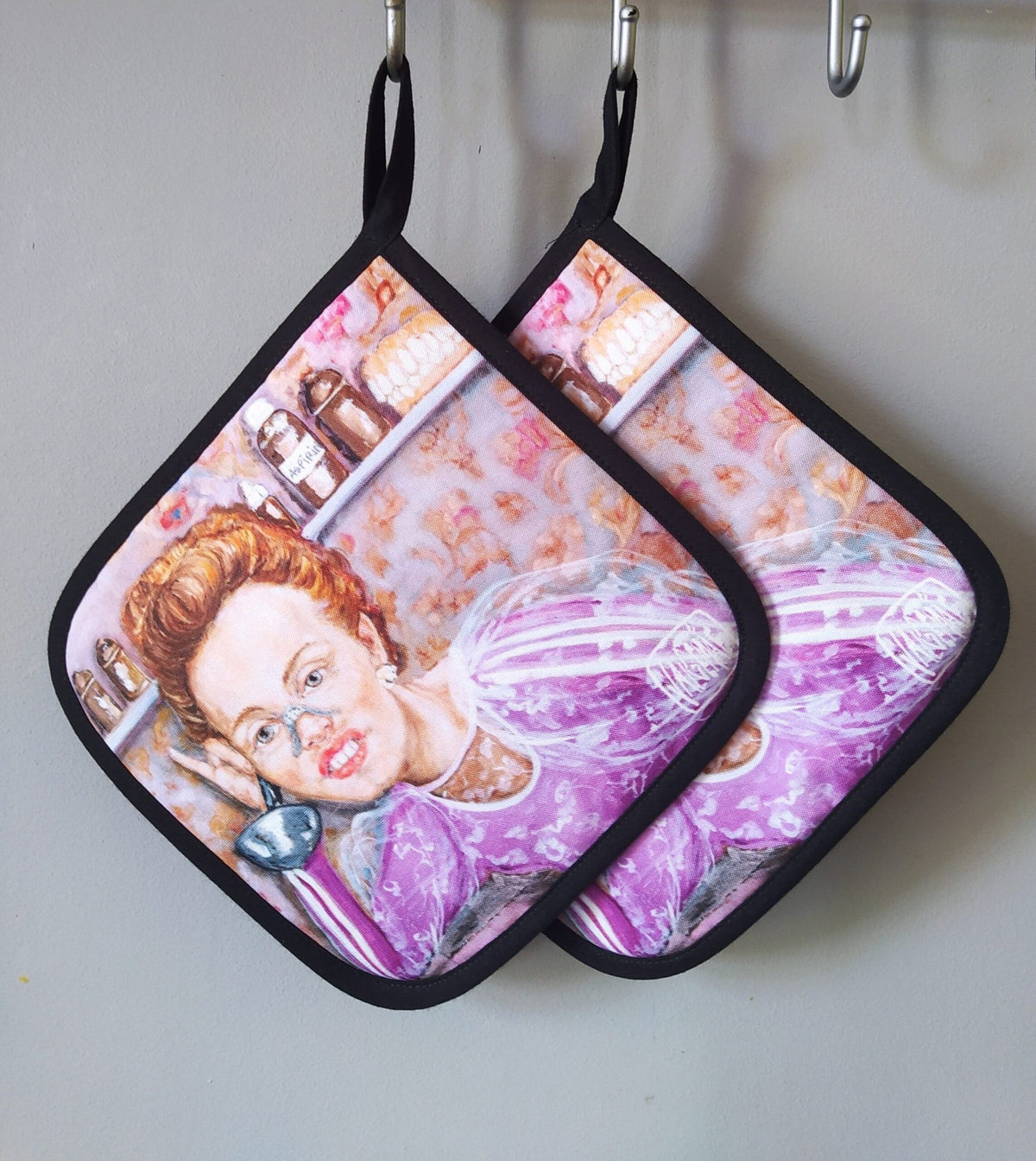 1. Pair of Katrin Valgemäe pot holders with pop art style painting of a woman in a pink dress, hanging on a kitchen wall