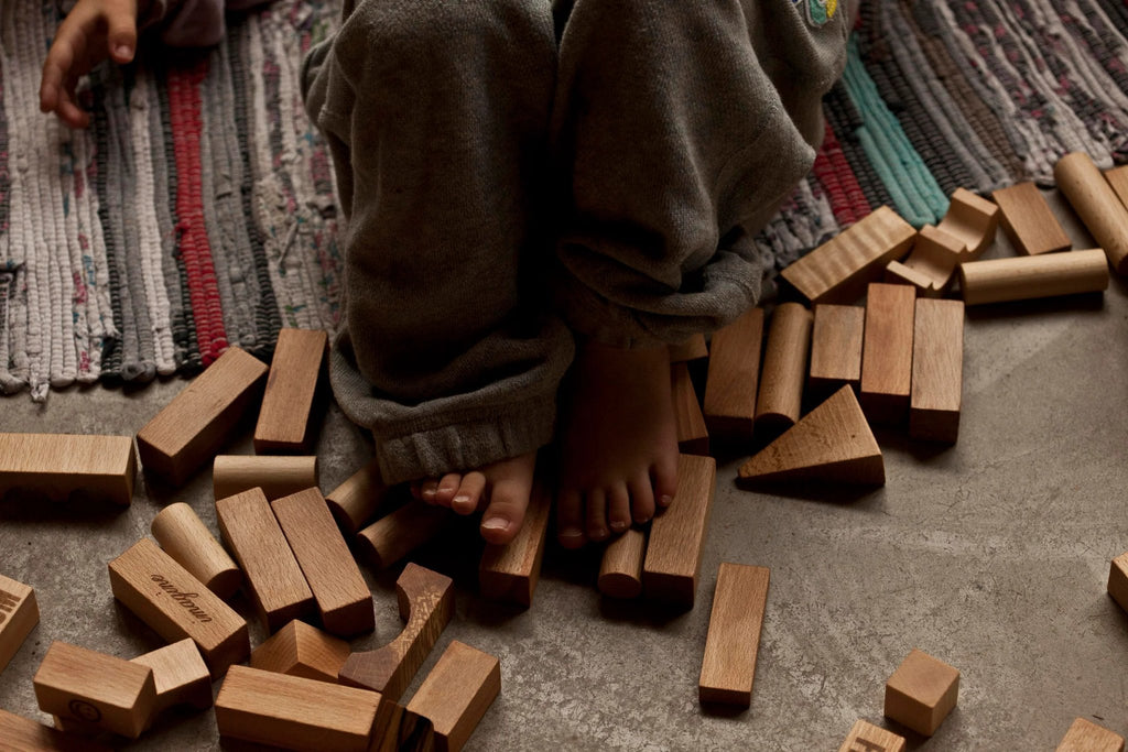 1. Child's feet among scattered Wooden Story natural wooden blocks on a textured rug and concrete floor