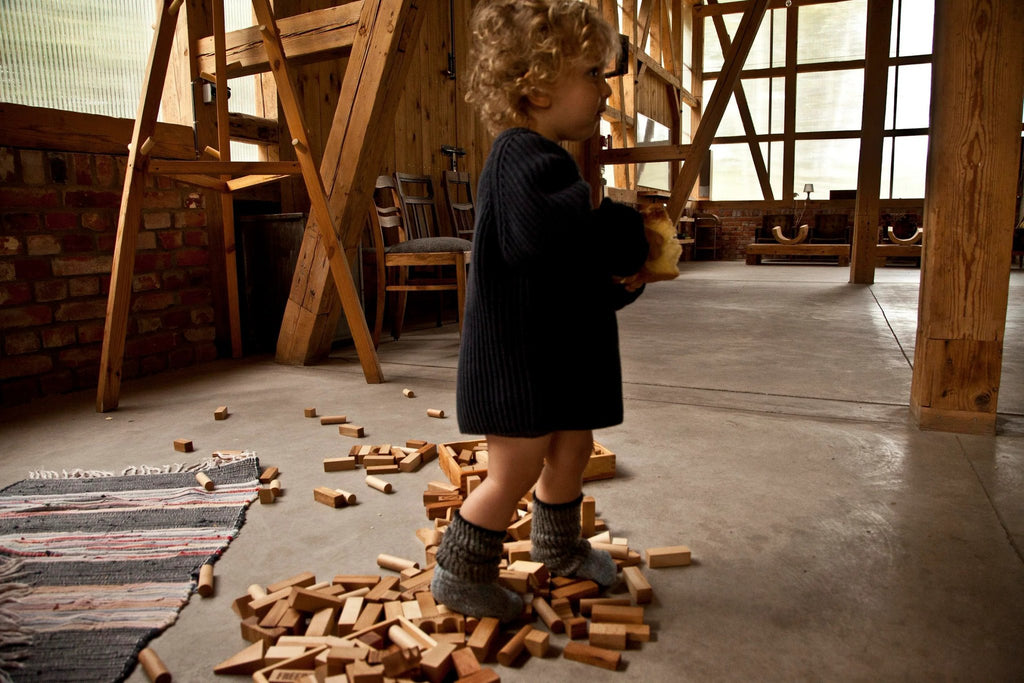 1. Child playing with Wooden Story natural wooden blocks in a rustic room, surrounded by scattered blocks on the floor