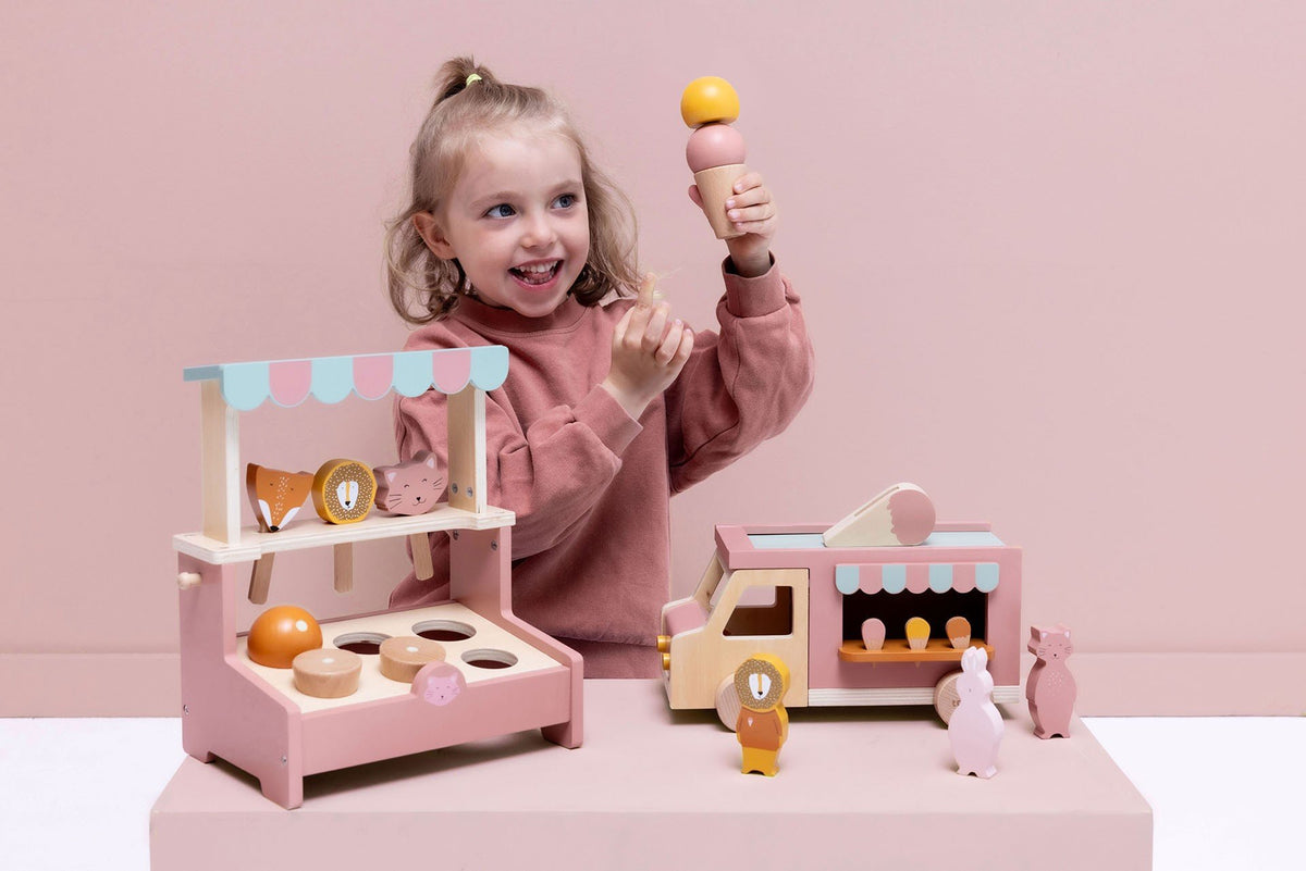 2. Smiling girl holding ice cream cone next to Trixie Baby wooden ice cream truck and stand, with animal figures on a pink background
