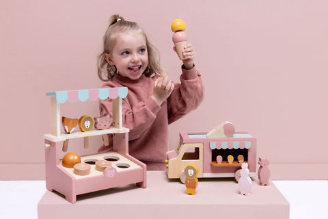 2. Smiling girl holding ice cream cone next to Trixie Baby wooden ice cream truck and stand, with animal figures on a pink background