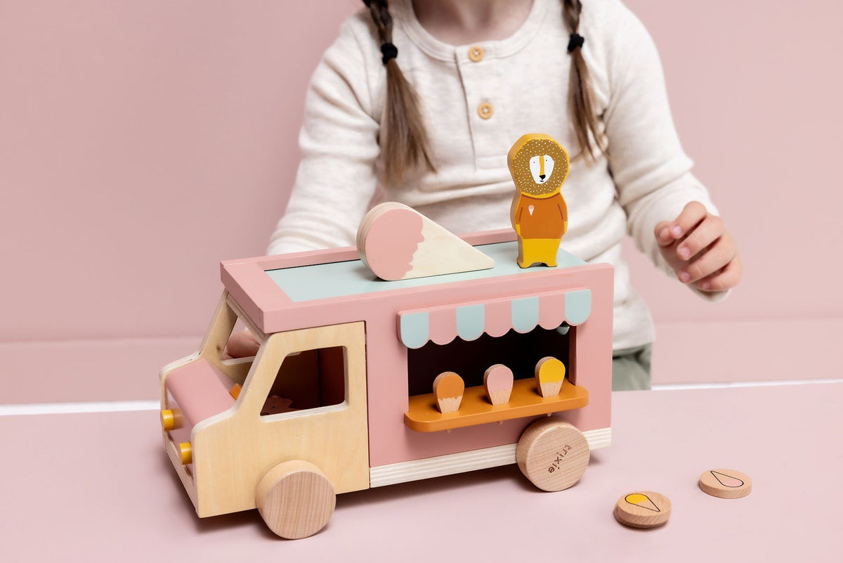 1. Child playing with Trixie Baby wooden ice cream truck featuring pink and natural wood colors, with animal figures and ice cream cones on a pink background