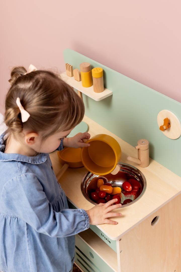 1. Young girl playing with Trixie Baby Wooden Kitchen Set in playroom, pouring from orange pot into sink