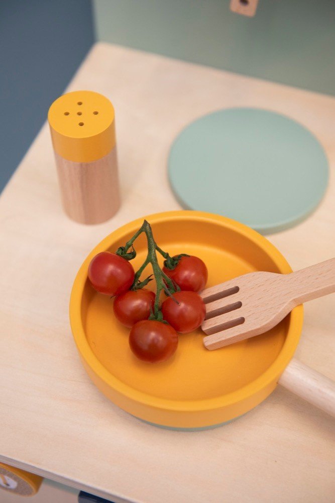 3. Close-up of Trixie Baby Mr. Lion wooden pan with cherry tomatoes and fork, next to a salt shaker on a play kitchen surface