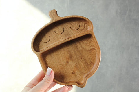 2. Person holding a wooden acorn-shaped plate for kids with two compartments against a grey background