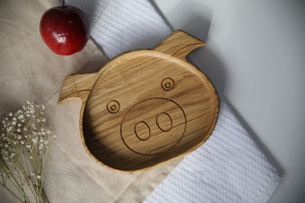 1. Handmade wooden boar-shaped plate for kids on a beige cloth with a red apple and white flowers, showcasing engraved face design