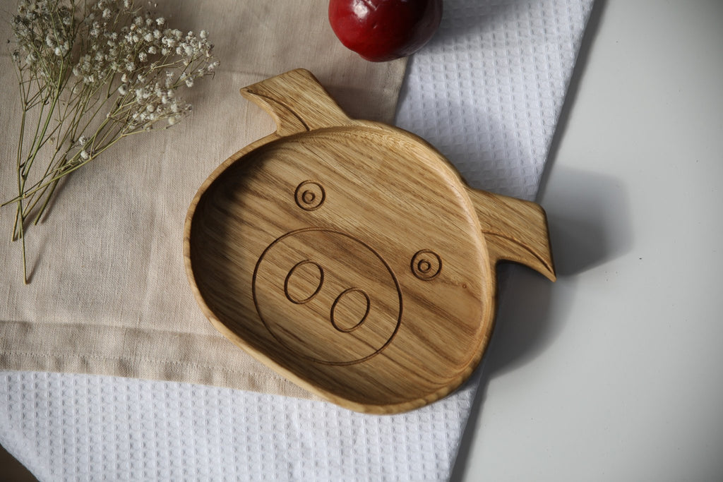 4. Front view of wooden boar-shaped plate for kids on a beige cloth with a red apple and white flowers, showing engraved face design