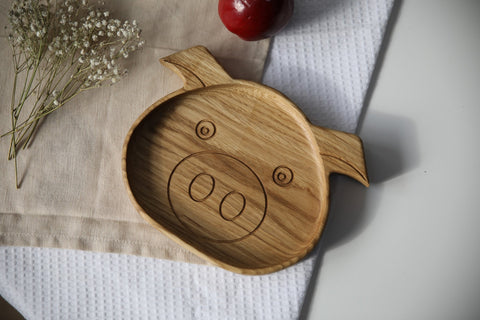 4. Front view of wooden boar-shaped plate for kids on a beige cloth with a red apple and white flowers, showing engraved face design
