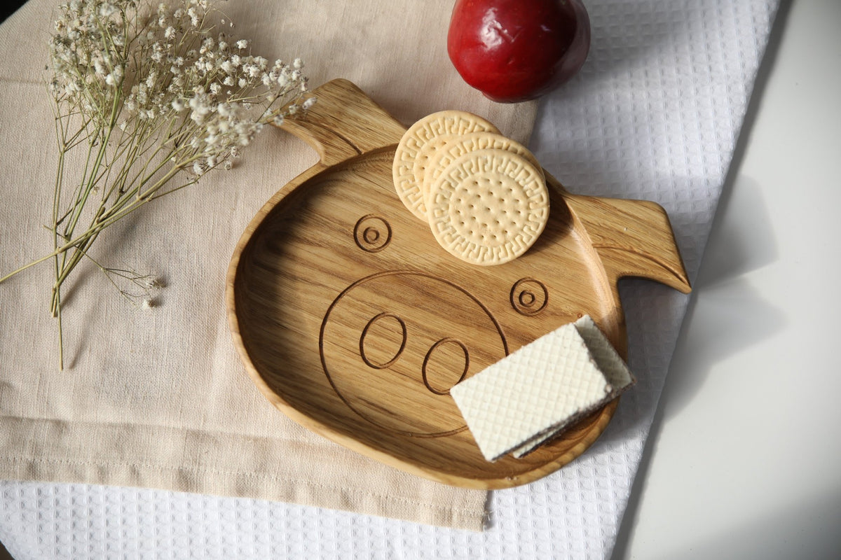 3. Wooden boar-shaped plate for kids with cookies and wafer on a beige cloth, accompanied by a red apple and white flowers