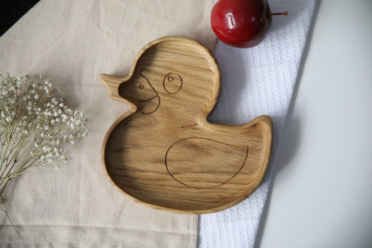 1. Handmade wooden duckling-shaped plate on a beige cloth with a red apple and white flowers, showcasing smooth oak or ash finish