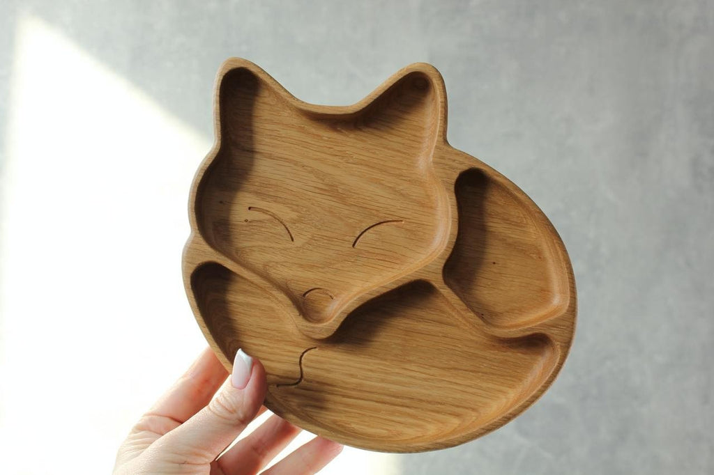 1. Person holding a wooden fox-shaped plate with three compartments against a grey background, highlighting the natural wood finish