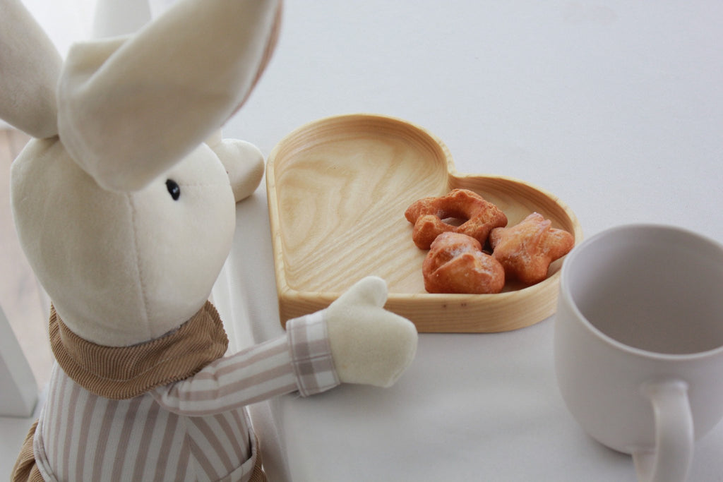 1. Plush toy rabbit sitting at table with heart-shaped wooden plate holding snacks, next to white mug
