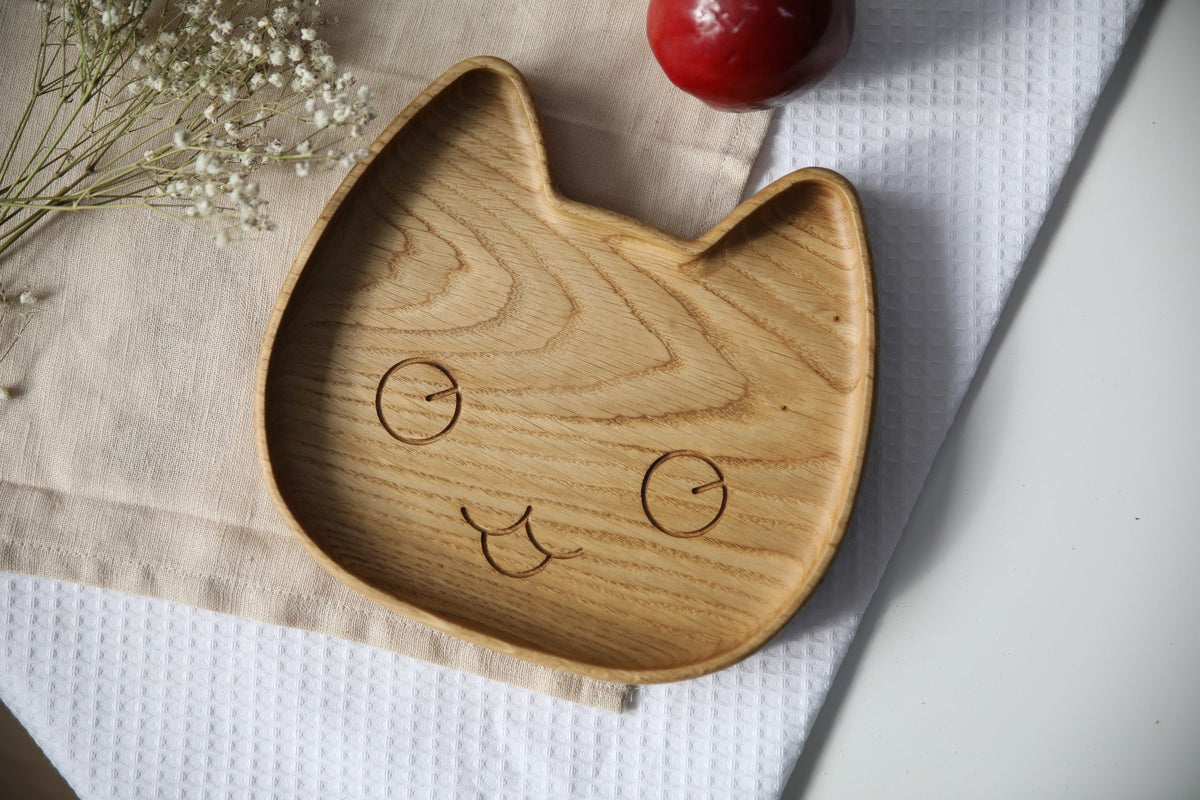 1. Handmade wooden plate for kids in cat shape on a textured cloth with flowers and an apple, showcasing smooth oak finish