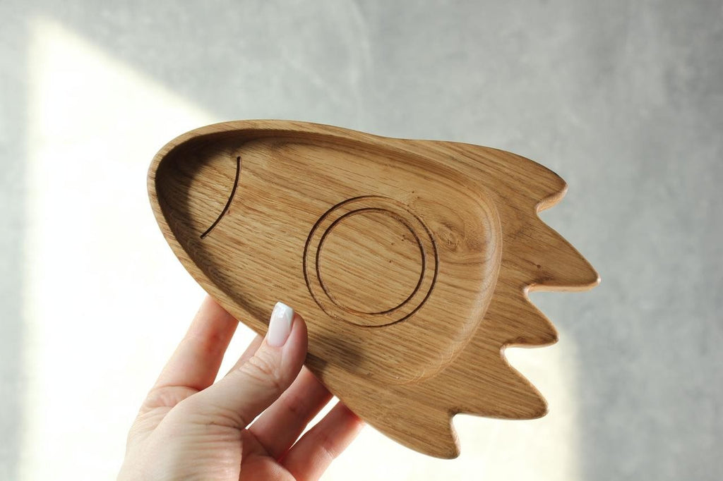 1. Hand holding a handmade wooden rocket-shaped plate for kids, made of oak or ash, against a light background