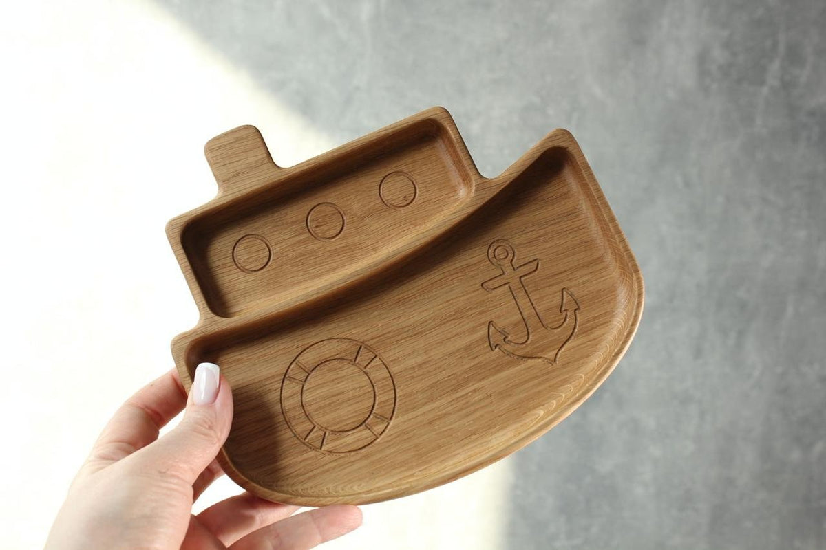 3. Hand holding a wooden ship-shaped plate for kids, featuring engraved anchor and lifebuoy, made from oak or ash, against a grey background