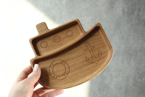 3. Hand holding a wooden ship-shaped plate for kids, featuring engraved anchor and lifebuoy, made from oak or ash, against a grey background