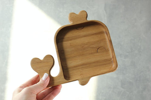 1. Person holding a handmade wooden whale-shaped plate for kids, showcasing its smooth, polished surface and natural wood grain against a light background.