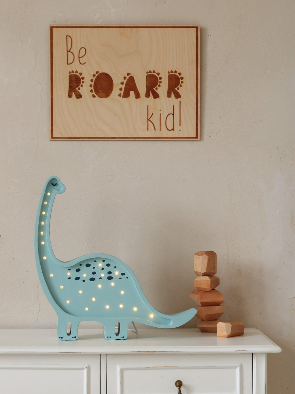 2. Engraved 'Be Roarr Kid' wooden poster on wall above white cabinet with blue dinosaur lamp and wooden blocks