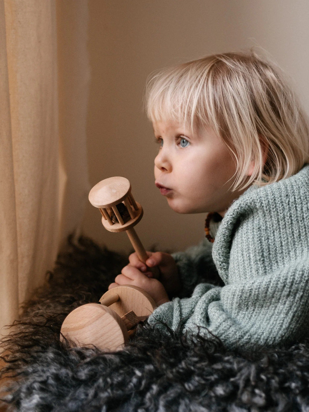 1. Child with blonde hair holding Wooden Story rattle while lying on a furry blanket near a window