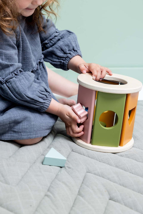 1. Child playing with Trixie Baby wooden rolling shape sorter on a quilted mat, inserting pink shape into pastel-colored sorter