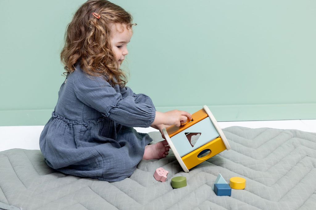 4. Child sitting on quilted mat playing with Trixie Baby wooden rolling shape sorter, surrounded by colorful shapes