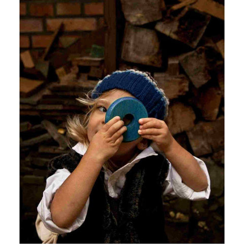3. Child holding a blue wooden disk from the round pyramid toy, wearing a blue knit hat and white shirt, with a woodpile in the background
