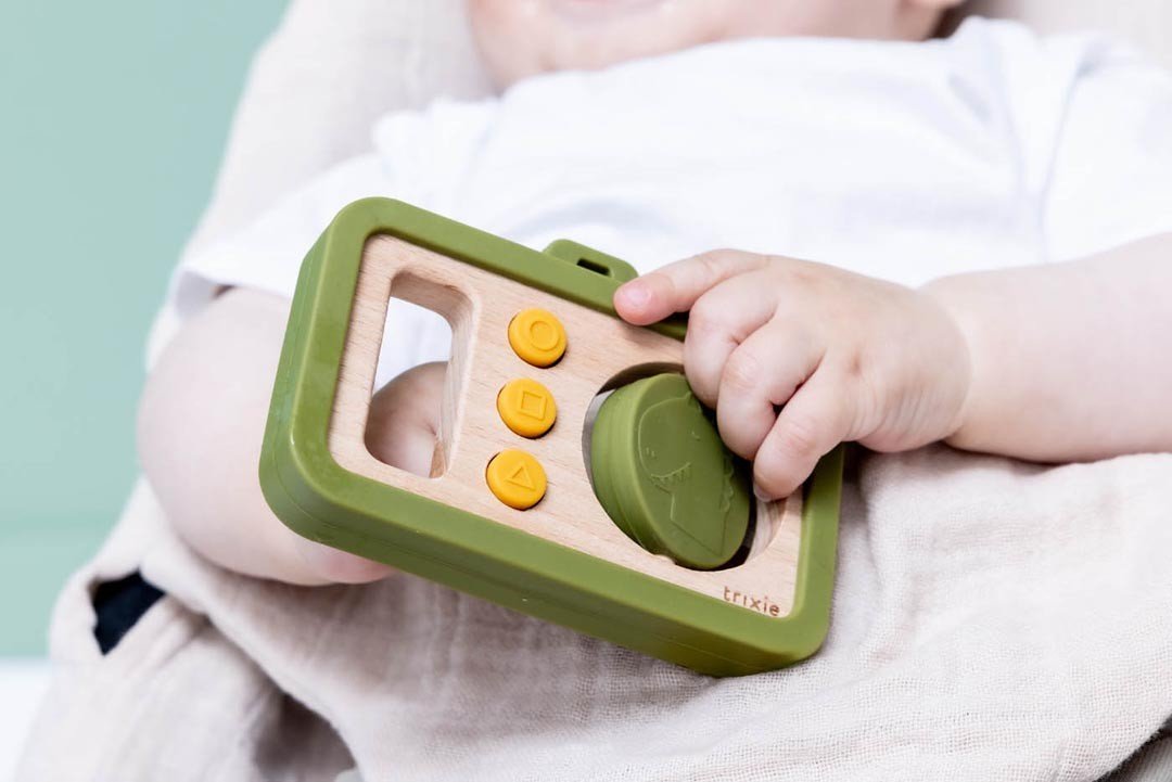 1. Baby holding Trixie Baby's wooden silicone camera with green frame and yellow buttons, showcasing easy grip and playful design
