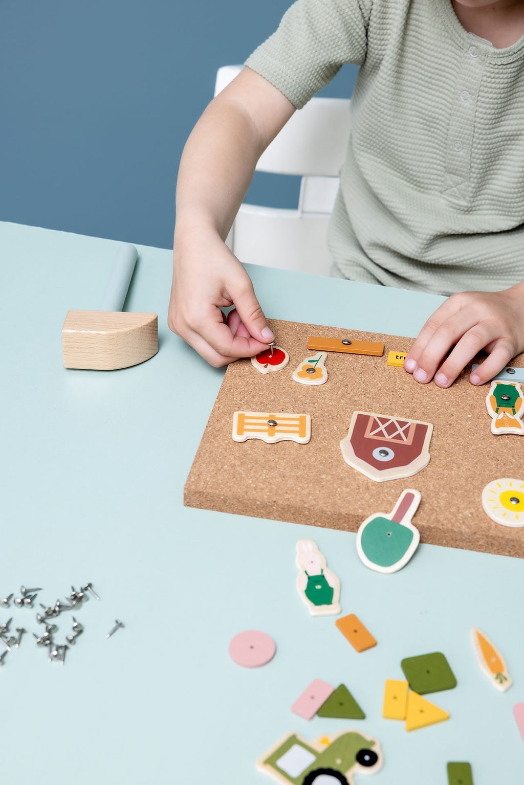 2. Close-up of child assembling Trixie Baby wooden tap tap game on cork board with farm-themed pieces, hammer visible on table