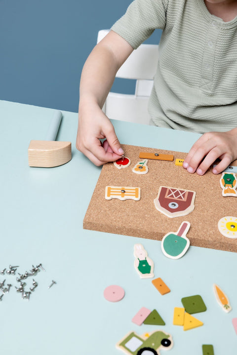 2. Close-up of child assembling Trixie Baby wooden tap tap game on cork board with farm-themed pieces, hammer visible on table