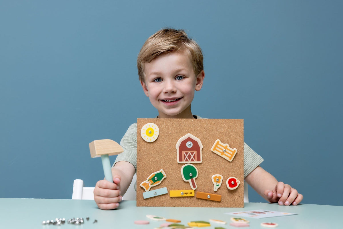 1. Child smiling while playing with Trixie Baby wooden tap tap game on cork board, featuring farmyard pieces like barn and animals, against blue background
