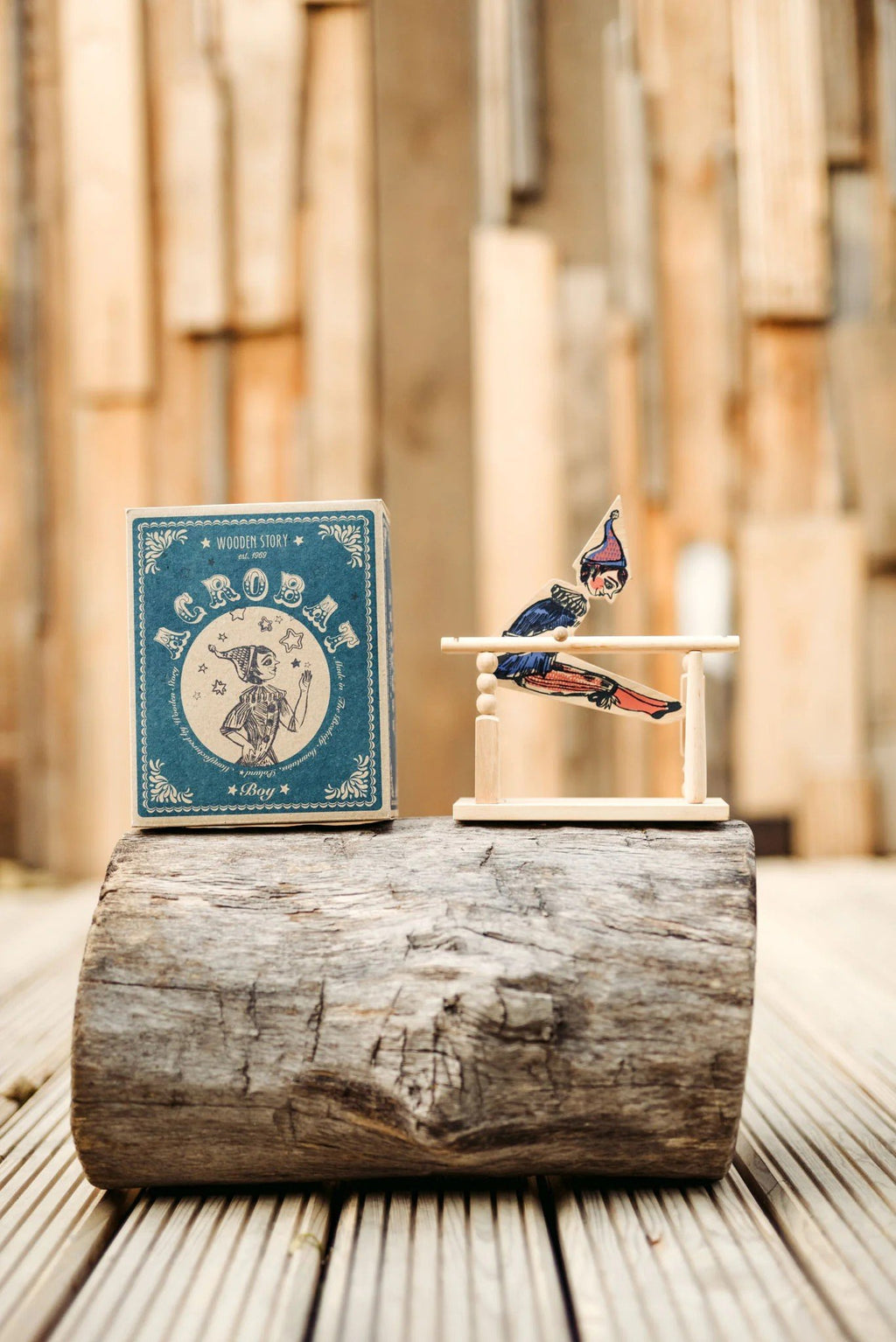 1. Wooden Story acrobat toy and box displayed on a wooden log with a rustic background
