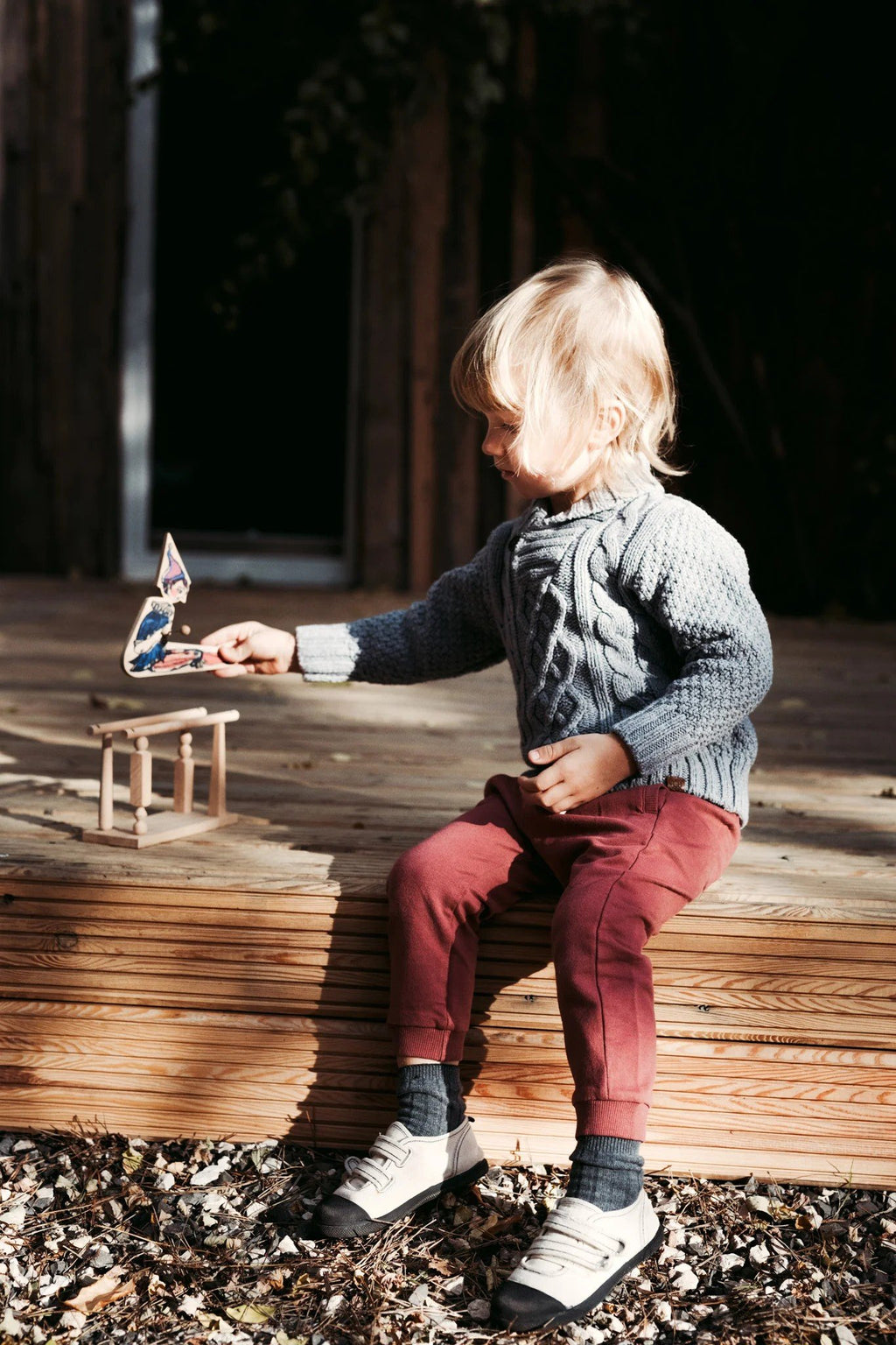 1. Child sitting on wooden steps playing with Wooden Story acrobat toy outdoors, wearing a grey sweater and red pants