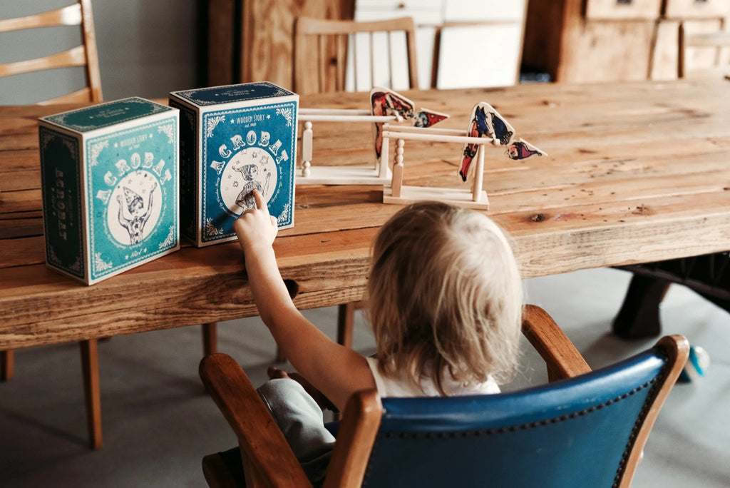 1. Child playing with Wooden Story acrobat toy on a wooden table, with two toy boxes in the background