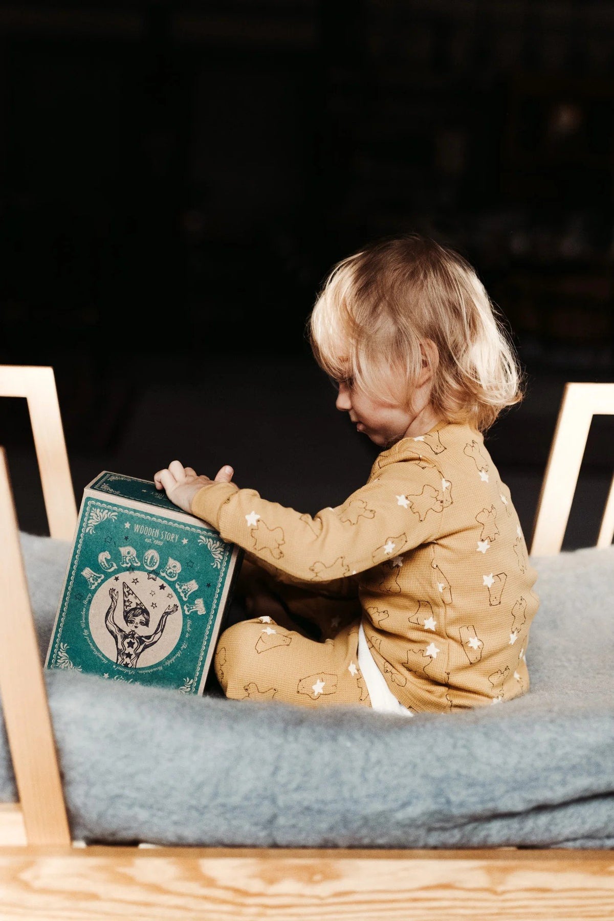 1. Child in yellow pajamas sitting on a bed, holding the Wooden Story acrobat toy box in a softly lit room