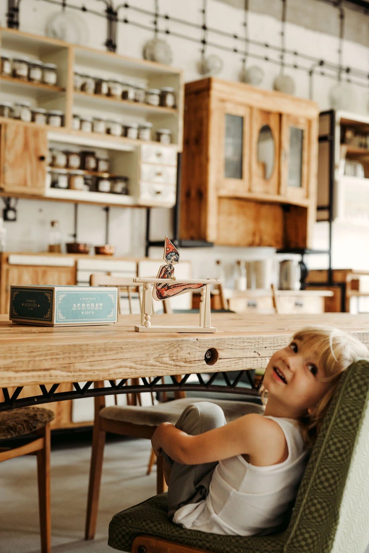 1. Smiling child sitting at a table with Wooden Story acrobat toy and packaging in a rustic kitchen setting