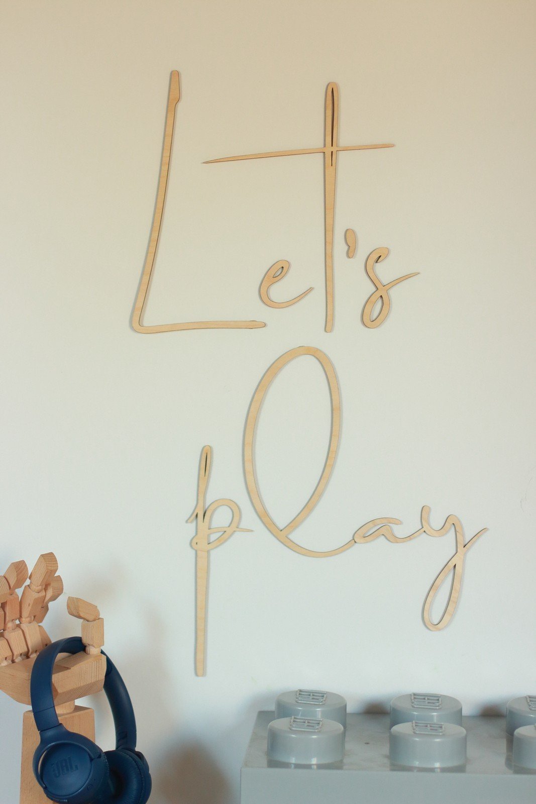 3. Natural plywood 'Let's Play' wall sign in children's play area with wooden toys and headphones on a shelf