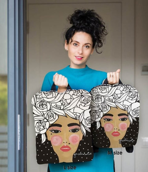 2. Woman in blue dress holding two sizes of Garbanota BACKPACK SADE with floral artwork, indoor setting